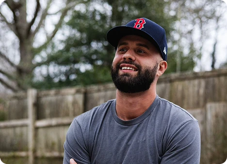 Smiling man with beard wearing a navy baseball cap and gray t-shirt outdoors beside a wooden fence.