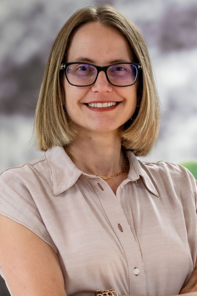 Smiling woman with shoulder-length blond hair and black-framed glasses wearing a striped button-up shirt and gold necklace.