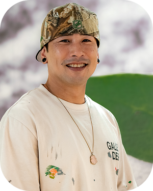 Smiling man wearing a camo bandana and beige paint-splattered shirt with a pendant necklace, standing outdoors.