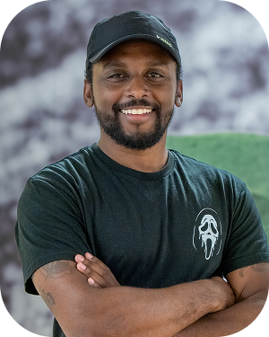 Smiling man with a cap standing outdoors with arms crossed, wearing a dark T-shirt with a small white logo.