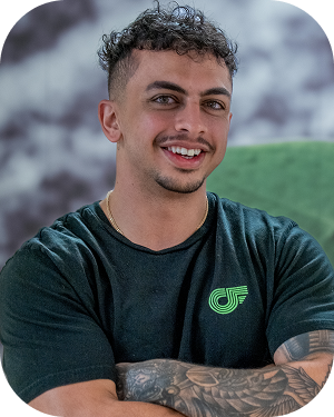 Smiling man with tattooed arms wearing a black t-shirt with a green logo, outdoor blurred background.