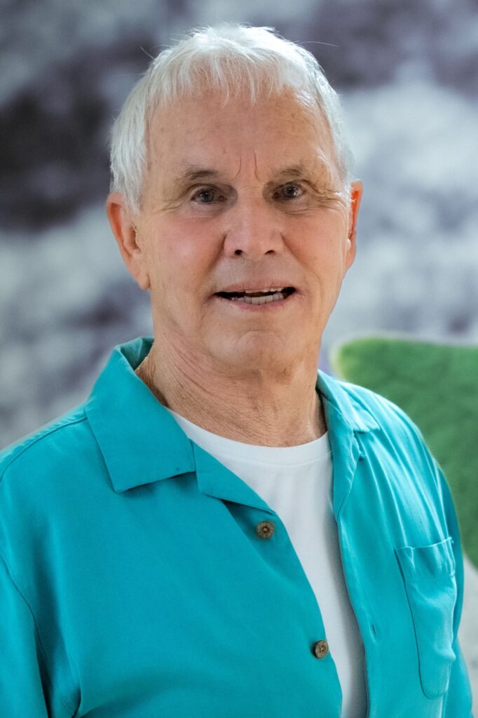 Close-up portrait of a smiling elderly man with white hair wearing a turquoise shirt, outdoors.