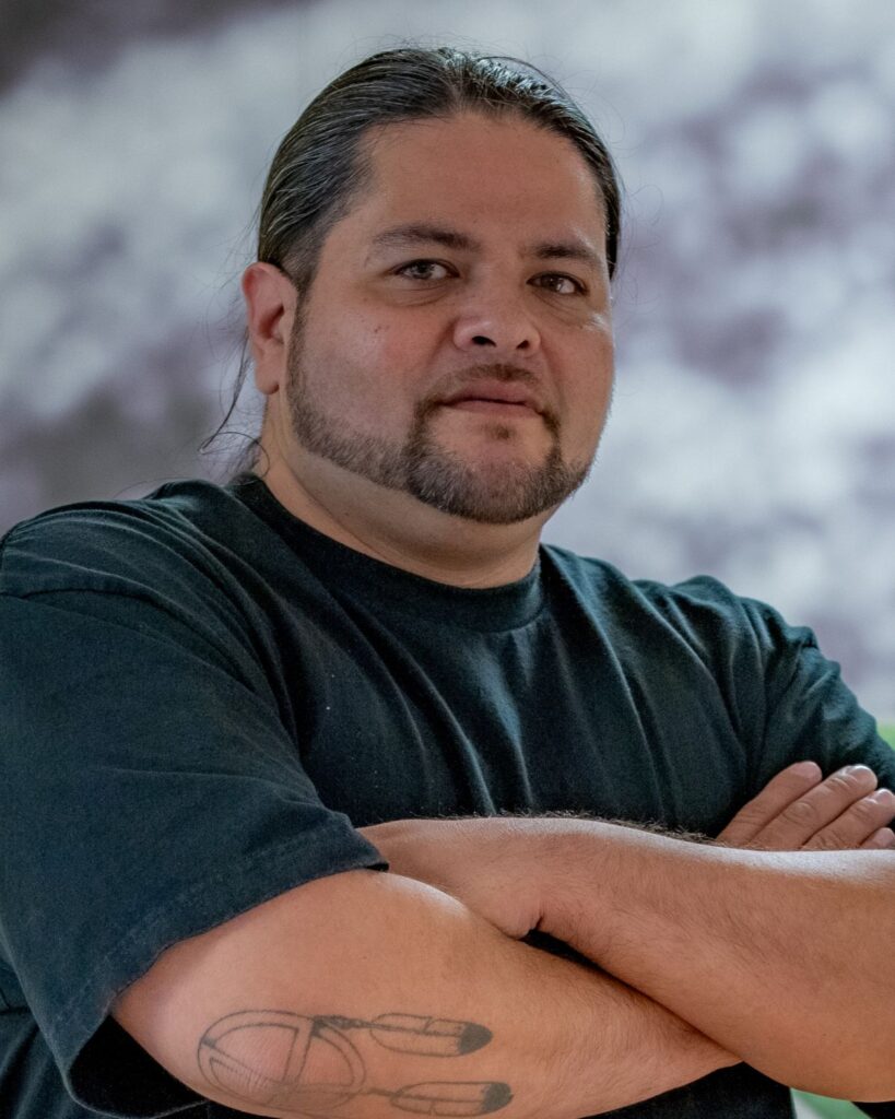 Man with long dark hair tied back, trimmed beard, and crossed arms wearing a black T-shirt; tattoo on forearm visible