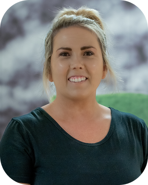 Smiling woman with blonde hair tied back, wearing a dark green shirt, posed for a portrait against a soft background.
