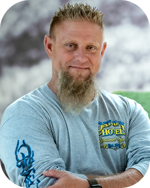 Portrait of a man with blond spiky hair and a goatee, wearing a light blue long-sleeve shirt, standing outdoors with arms crossed.