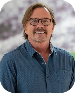 Smiling man with light brown hair and a goatee, wearing round glasses and a blue button-down shirt, outdoors on a blurred background.