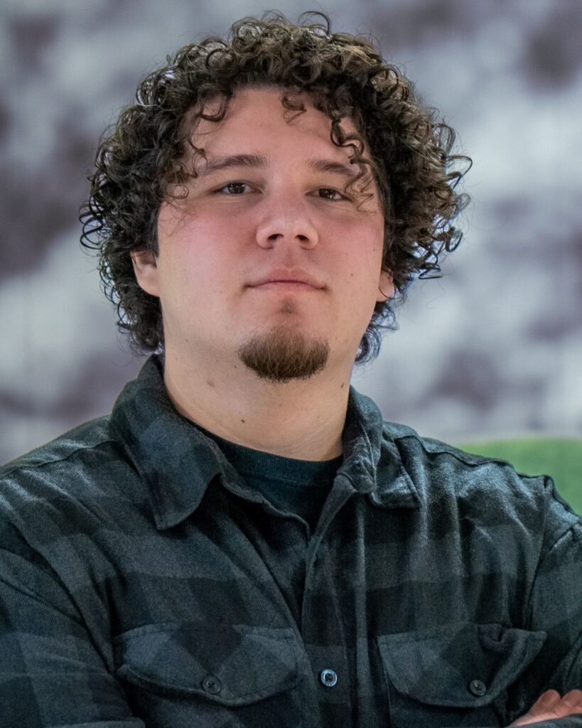 Portrait of a man with curly brown hair and a goatee, wearing a dark plaid shirt and crossing his arms.