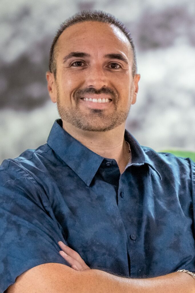 Smiling man with short dark hair and stubble, wearing a blue button-down shirt with his arms crossed (portrait).