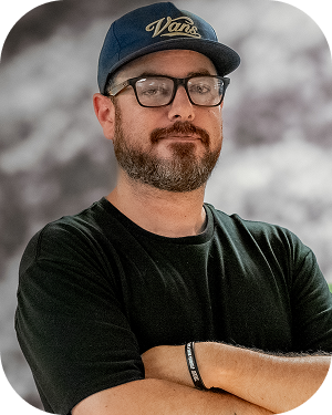 Man with glasses and a beard wearing a blue cap and black t-shirt, arms crossed, outdoors on a snowy day.