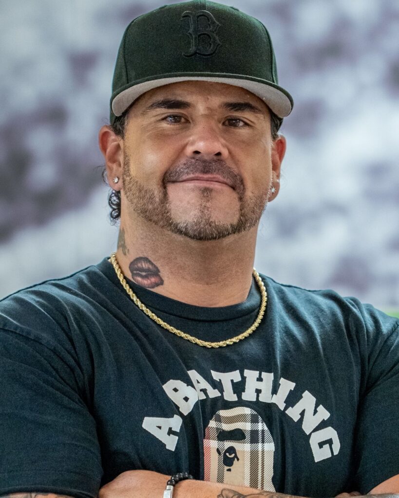 Portrait of a man wearing a dark cap, goatee, earrings, and a gold chain, with a neck tattoo and a black graphic T‑shirt.