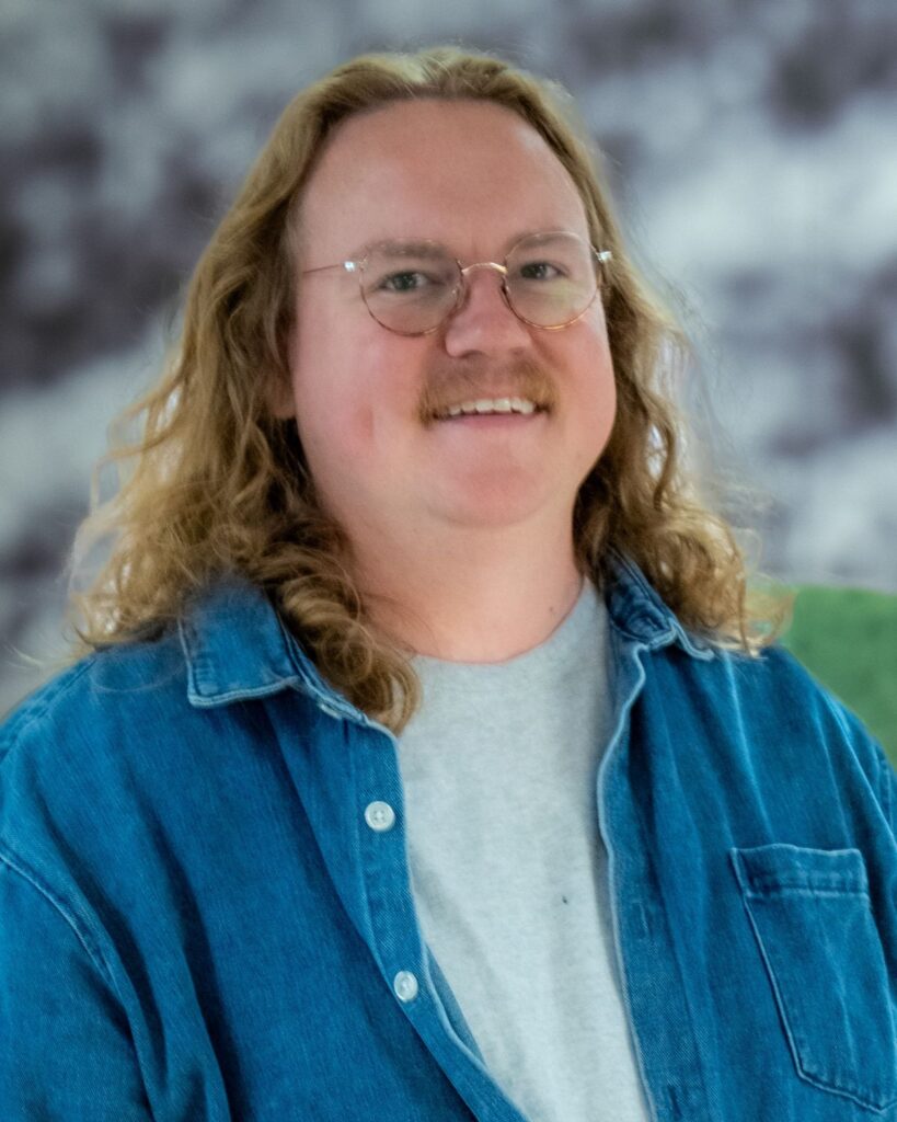 Person with shoulder-length wavy blond hair, round glasses, and a blue denim shirt, smiling.